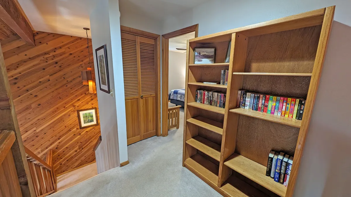 A cozy upstairs landing featuring a bookshelf filled with books, wood paneling, and a staircase leading down. The space is softly lit and inviting, offering a glimpse into a tranquil reading nook perfect for quiet moments after a day of exploration.