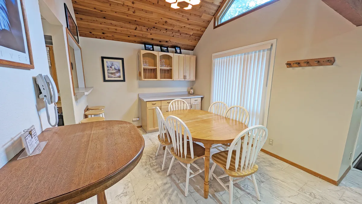 A cozy dining area with a wooden table and white chairs, set near cabinets with glass doors and a sliding glass door that hints at easy indoor-outdoor flow. The setup is primed for shared meals, board games, or morning coffee gatherings.