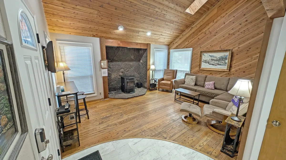 A welcoming living room featuring a stone fireplace, wooden ceiling, comfortable seating area, and large windows looking out toward the outdoors. The space is ideal for relaxing after an adventurous day, gathering with family for movie nights, or simply enjoying a tranquil evening by the fire.