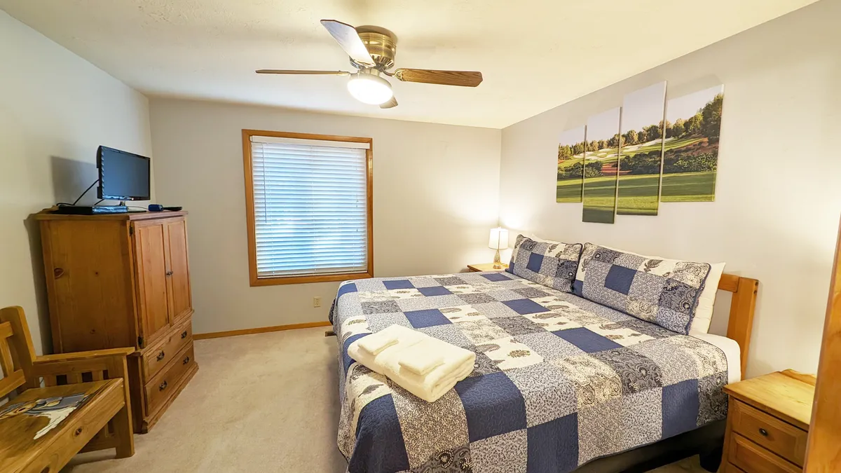 A cozy and relaxed primary suite, this room includes a spacious bed with a patterned duvet, wooden furniture, landscape artwork, and a television. The soft natural light and tranquil decor create an inviting space for relaxation and privacy.
