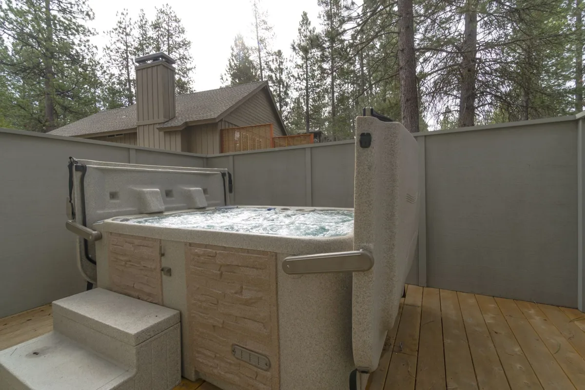 Shows a hot tub nestled beside privacy fencing and surrounded by mature trees, highlighting both relaxation and seclusion. The tranquil mood suggests morning soaks or quiet moments with coffee, as well as evening stargazing.