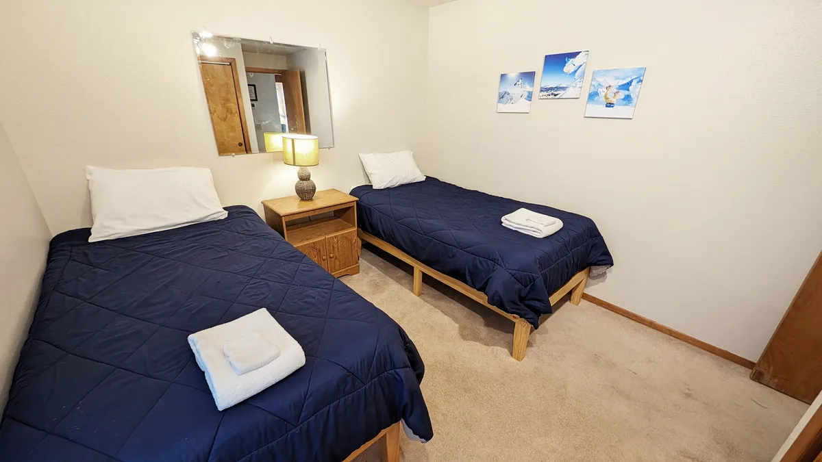 This image presents a downstairs bedroom featuring two twin beds separated by a nightstand with a lamp. The decor is simple and serene, with soft lighting and decorative wall photos, creating an inviting atmosphere for kids, friends, or solo travelers.
