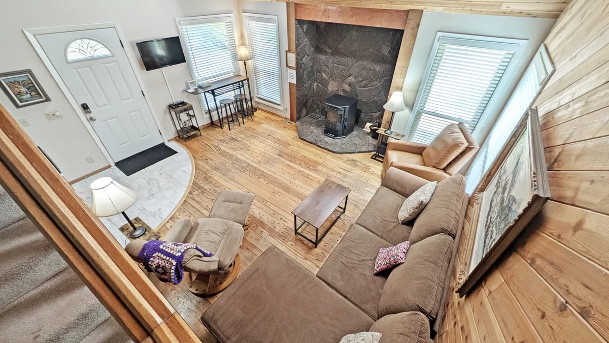 The image showcases the warm and inviting living room of a classic Sunriver cabin, featuring a glowing pellet stove fireplace, plush sofa, gleaming hardwood floors, and expansive windows that fill the space with natural light. The cozy arrangement invites relaxation and togetherness, perfect for unwinding after a day of adventure.