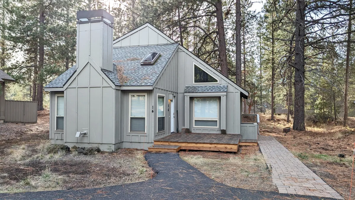 This image shows the exterior of the cabin featuring a spacious wooden deck, large windows, a prominent chimney, and a sloped roof, all set against a backdrop of pine trees. The setting hints at easy access to nature and a relaxed atmosphere that invites both exploration and downtime.