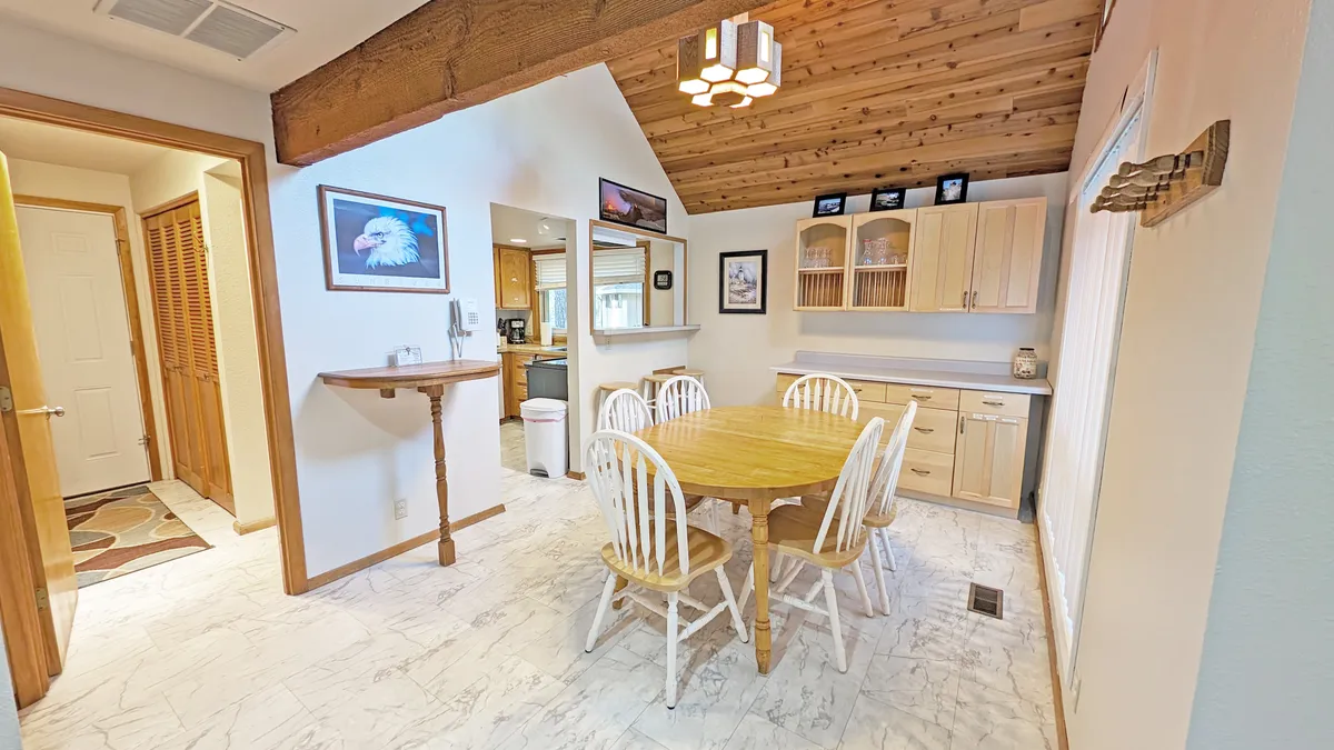A warm, inviting space featuring a large wooden dining table, kitchen cabinetry, wooden beams, and tasteful artwork. The open flow between the dining area and kitchen is evident, emphasizing the connection between cooking and gathering.