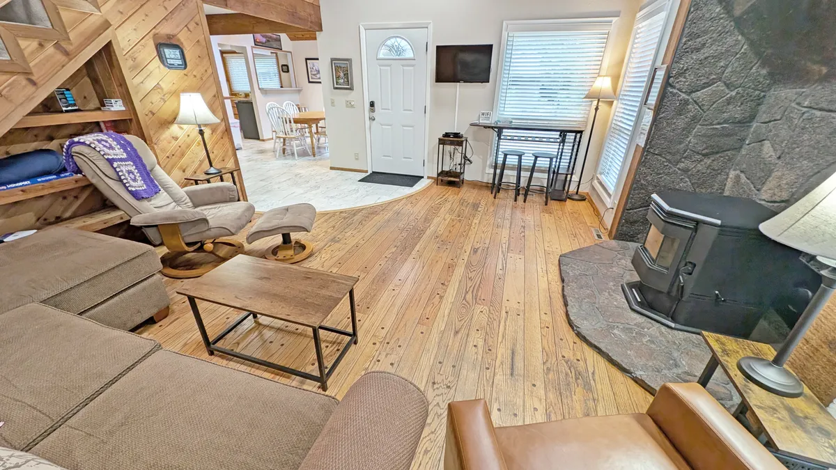 This image showcases a spacious, open-concept living area with a stone fireplace, comfortable seating, wooden floors, and a dining space visible in the background. The room is illuminated by natural light streaming in from the windows, highlighting the vaulted ceilings and rich wood accents.