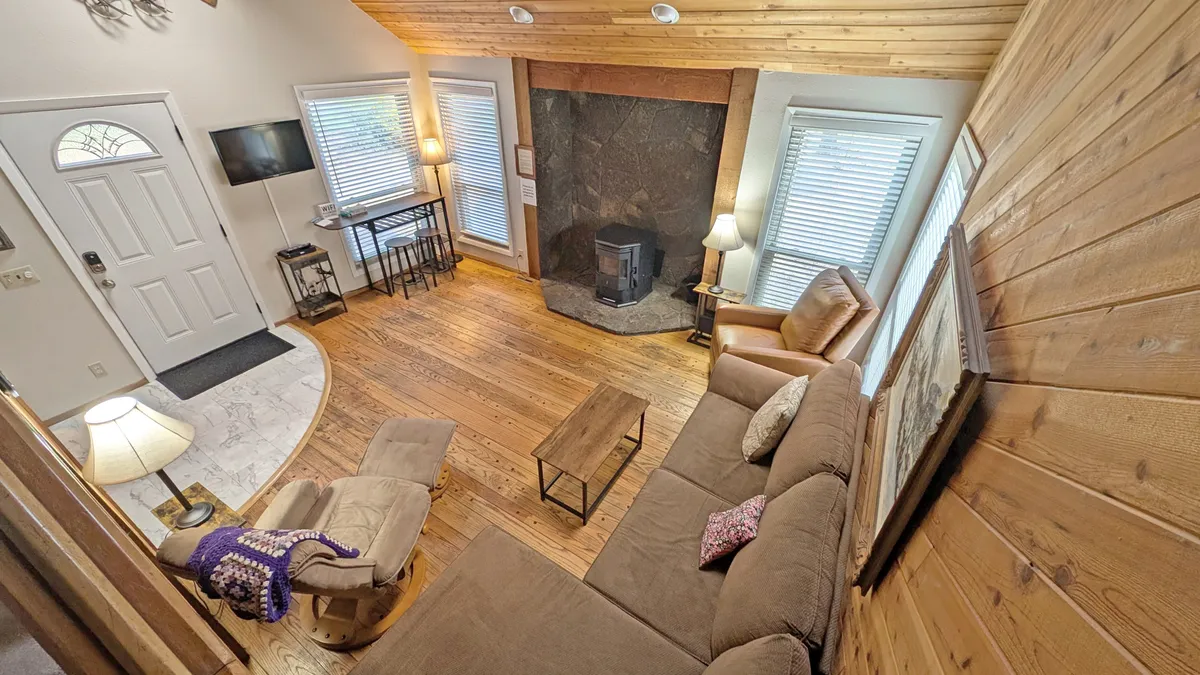 This image shows an inviting living room with a cozy fireplace, comfortable seating area, a front door that visually connects the interior space to the outdoors, and wooden flooring. Natural light is present, suggesting proximity to the outside environment.